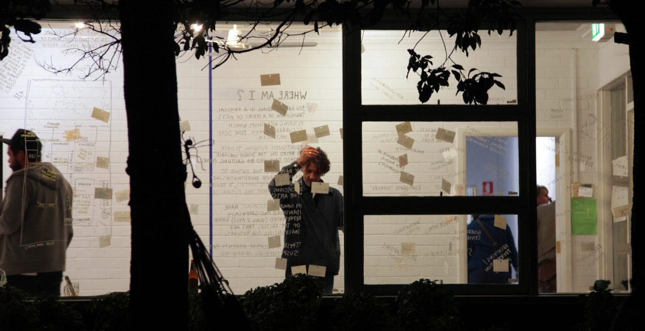 Photograph of the front room of Frontyard. Cards are suspended from the roof across a room, and people look at them, thinking about what the future described might be like.